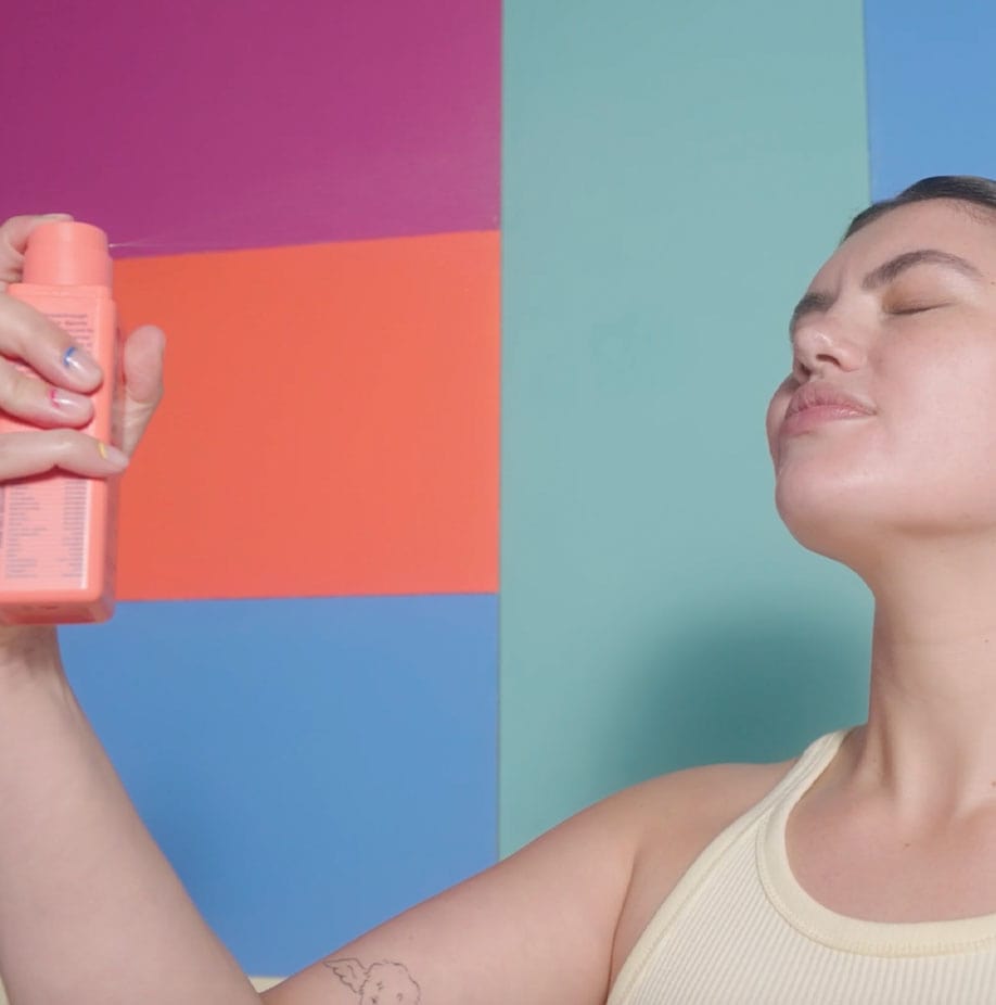 Woman spraying face mist with eyes closed against a colorful background.