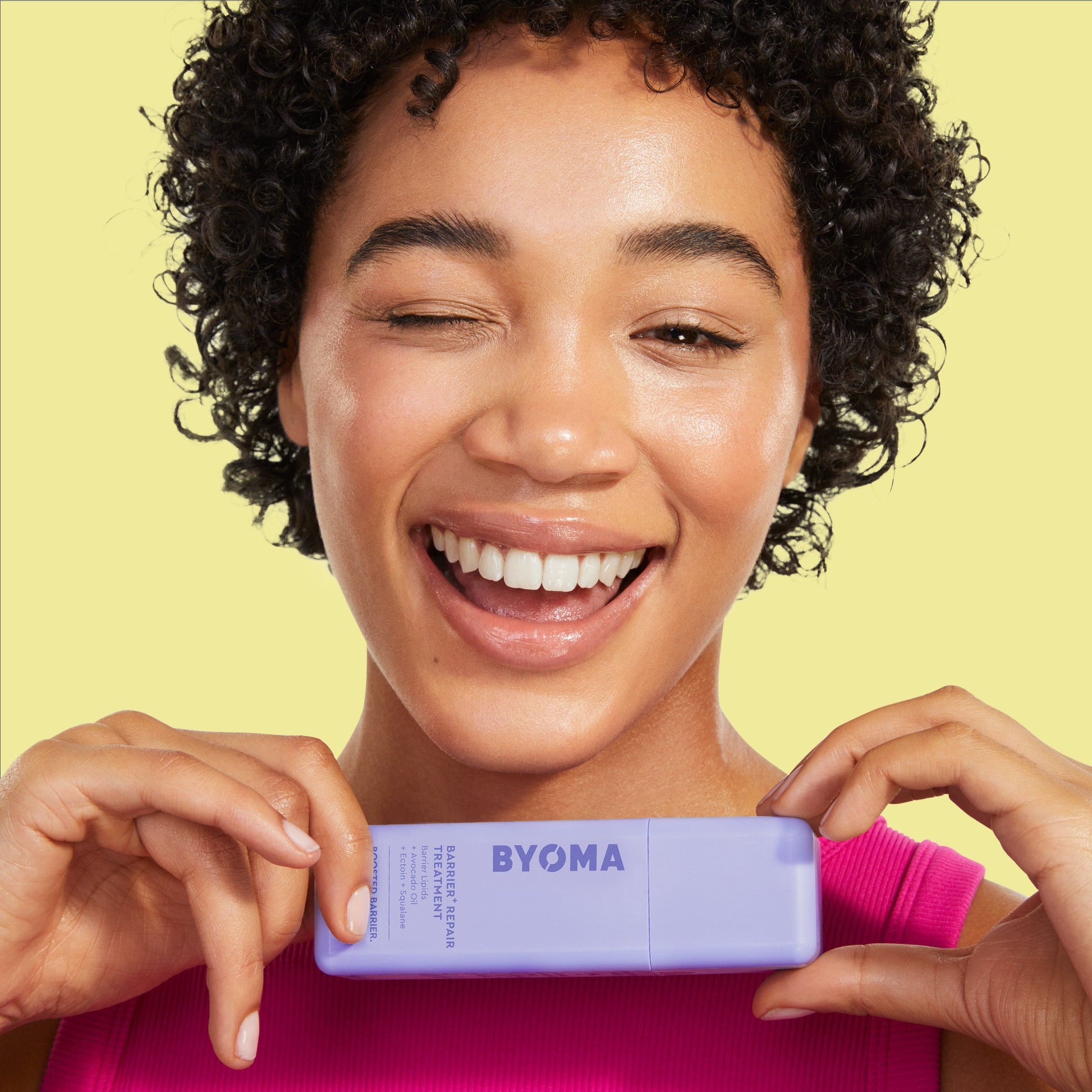 A woman with curly hair smiles and winks while holding Byoma Barrier Repair Treatment bottle.
